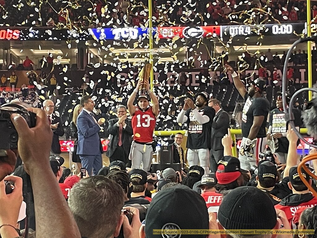 A football player holds up the championship trophy as confetti falls during a celebration at a packed stadium. Teammates and officials stand nearby, while fans cheer in the background.