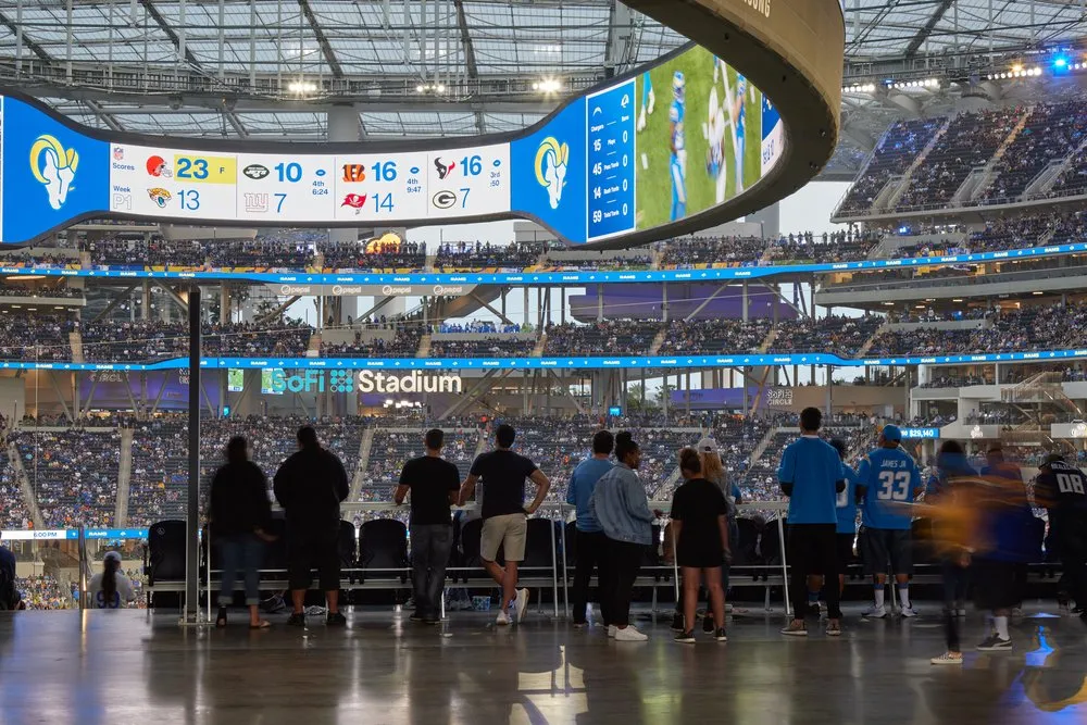 Fans inside SoFi Stadium watching a football game, with the large oval video board displaying scores and game highlights. The stadium is filled with spectators across multiple levels.