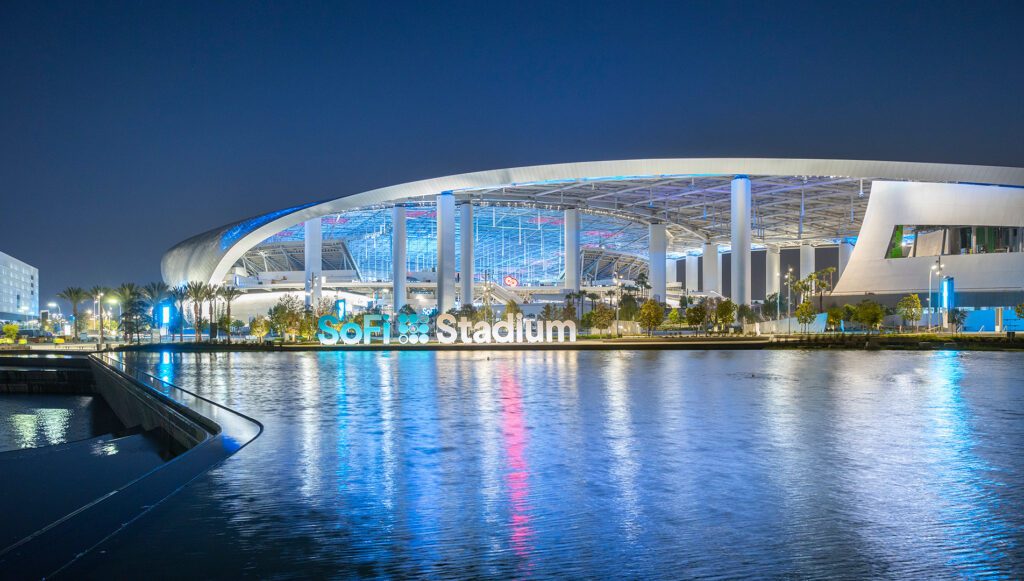 A nighttime view of SoFi Stadium, illuminated with blue and white lights, reflecting on the water in the foreground. The modern architectural design of the stadium is highlighted against the dark sky.