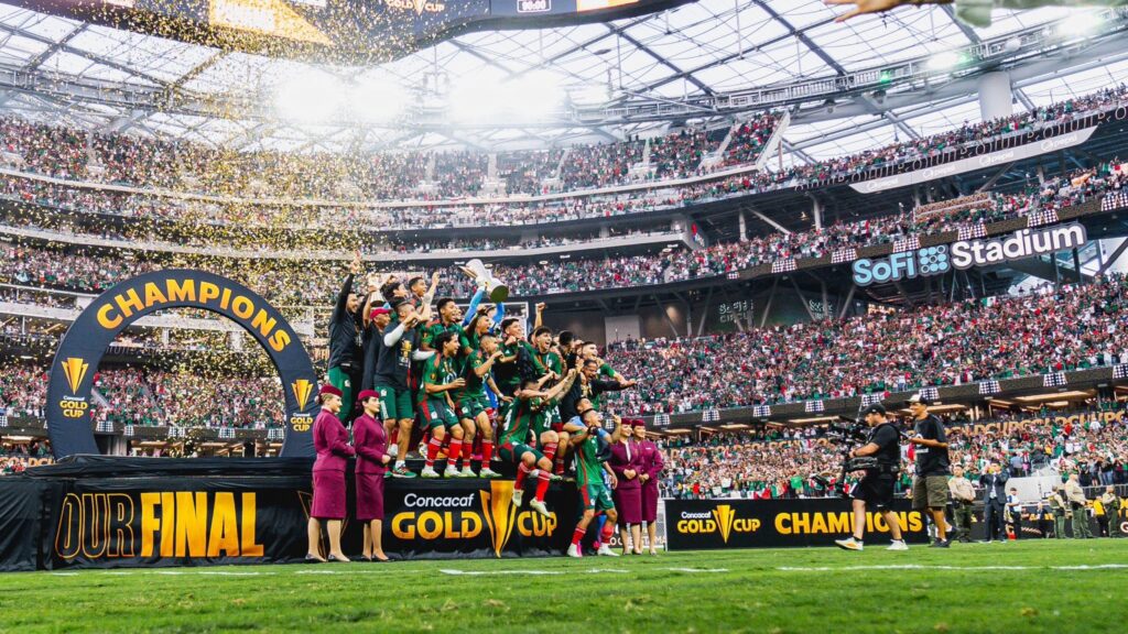 A triumphant soccer team celebrates winning the Concacaf Gold Cup at SoFi Stadium. The players stand on a stage under a large "Champions" arch while confetti rains down, surrounded by a cheering crowd in the stadium.