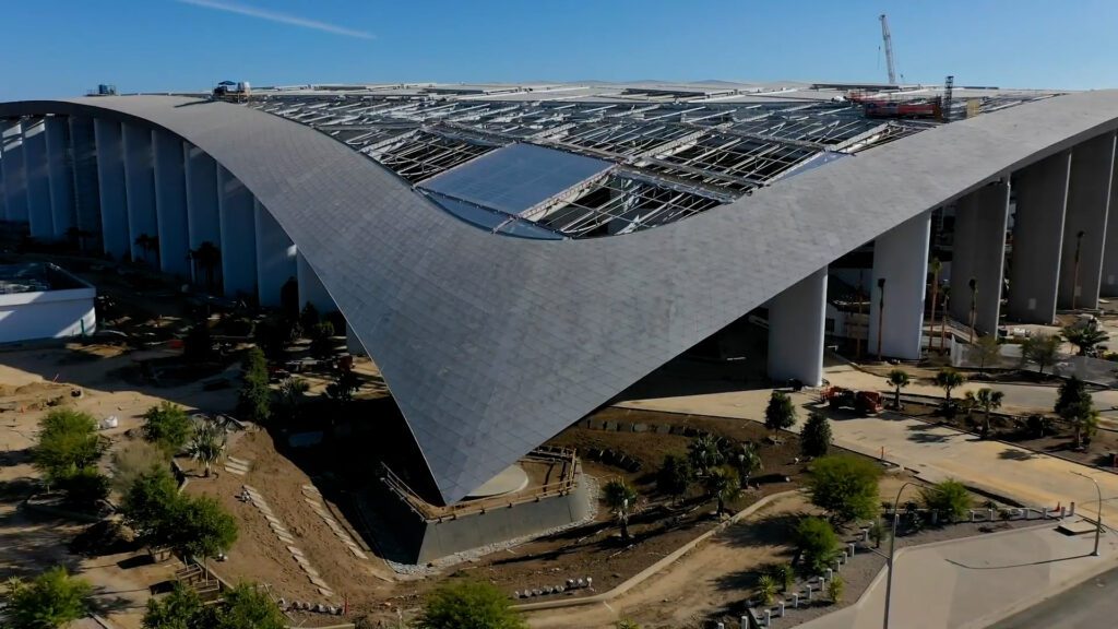 A close-up aerial view of SoFi Stadium during its construction, showcasing the curved roof structure, open framework, and surrounding landscaping under development.