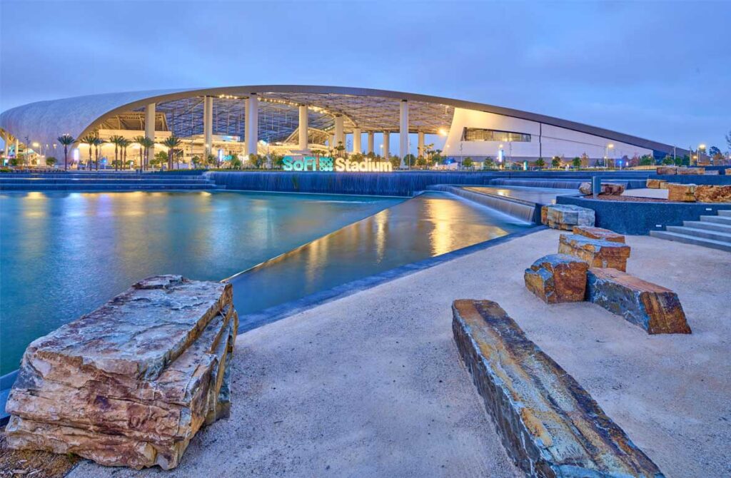 A scenic evening view of SoFi Stadium, illuminated with lights reflecting off the water in the foreground. The modern architecture is complemented by landscaped surroundings and a stone seating area.