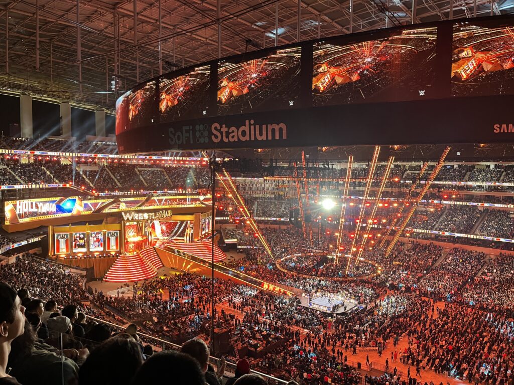 A vibrant view of a wrestling event at SoFi Stadium, with a packed crowd and colorful stage lighting. The large video board displays the event, and the arena is filled with excitement and energy.