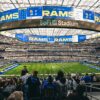 A wide-angle view of a football game at SoFi Stadium, with fans in the foreground wearing Los Angeles Rams jerseys. The massive scoreboard displays "Rams" and game details, while the field and seating areas are filled with spectators.