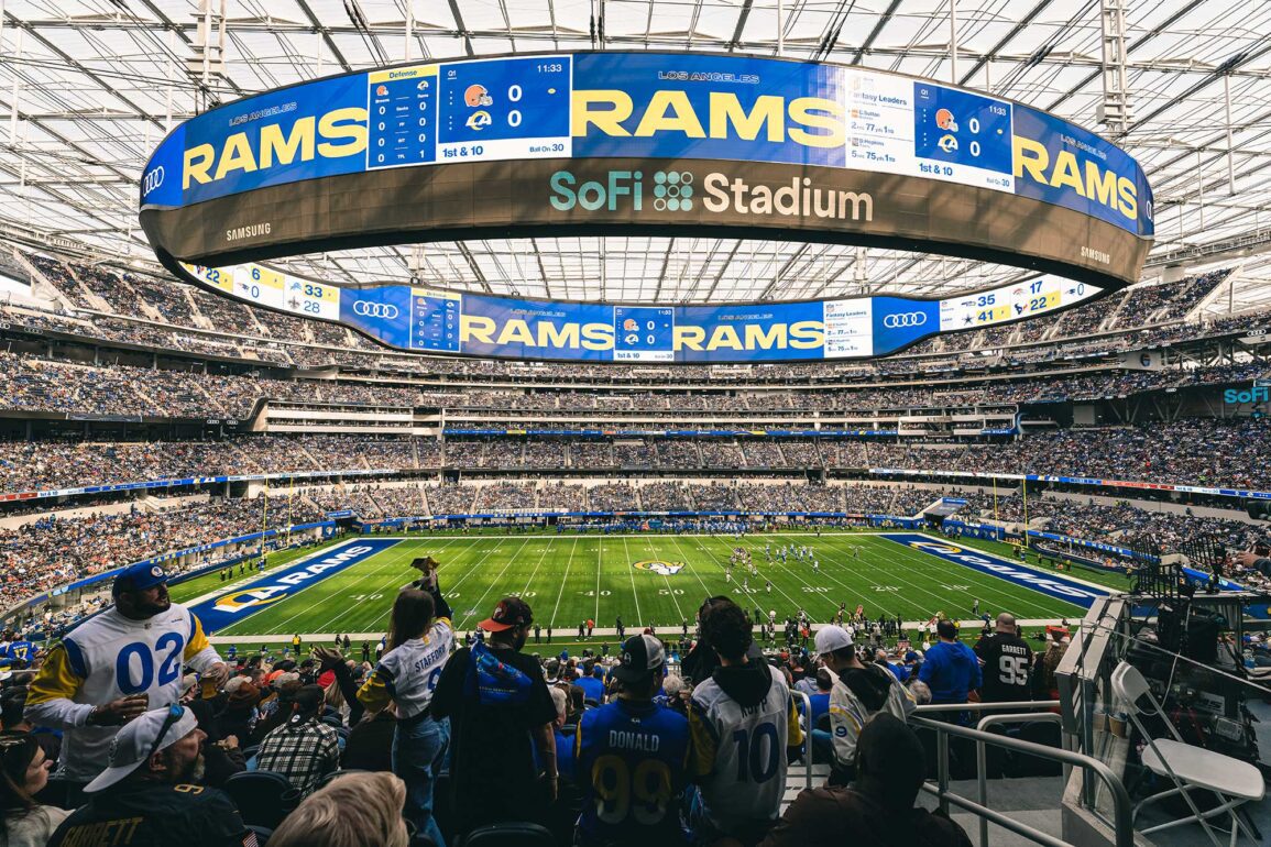 A wide-angle view of a football game at SoFi Stadium, with fans in the foreground wearing Los Angeles Rams jerseys. The massive scoreboard displays "Rams" and game details, while the field and seating areas are filled with spectators.