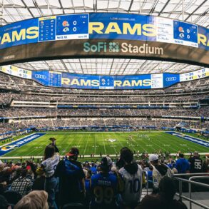 A wide-angle view of a football game at SoFi Stadium, with fans in the foreground wearing Los Angeles Rams jerseys. The massive scoreboard displays "Rams" and game details, while the field and seating areas are filled with spectators.